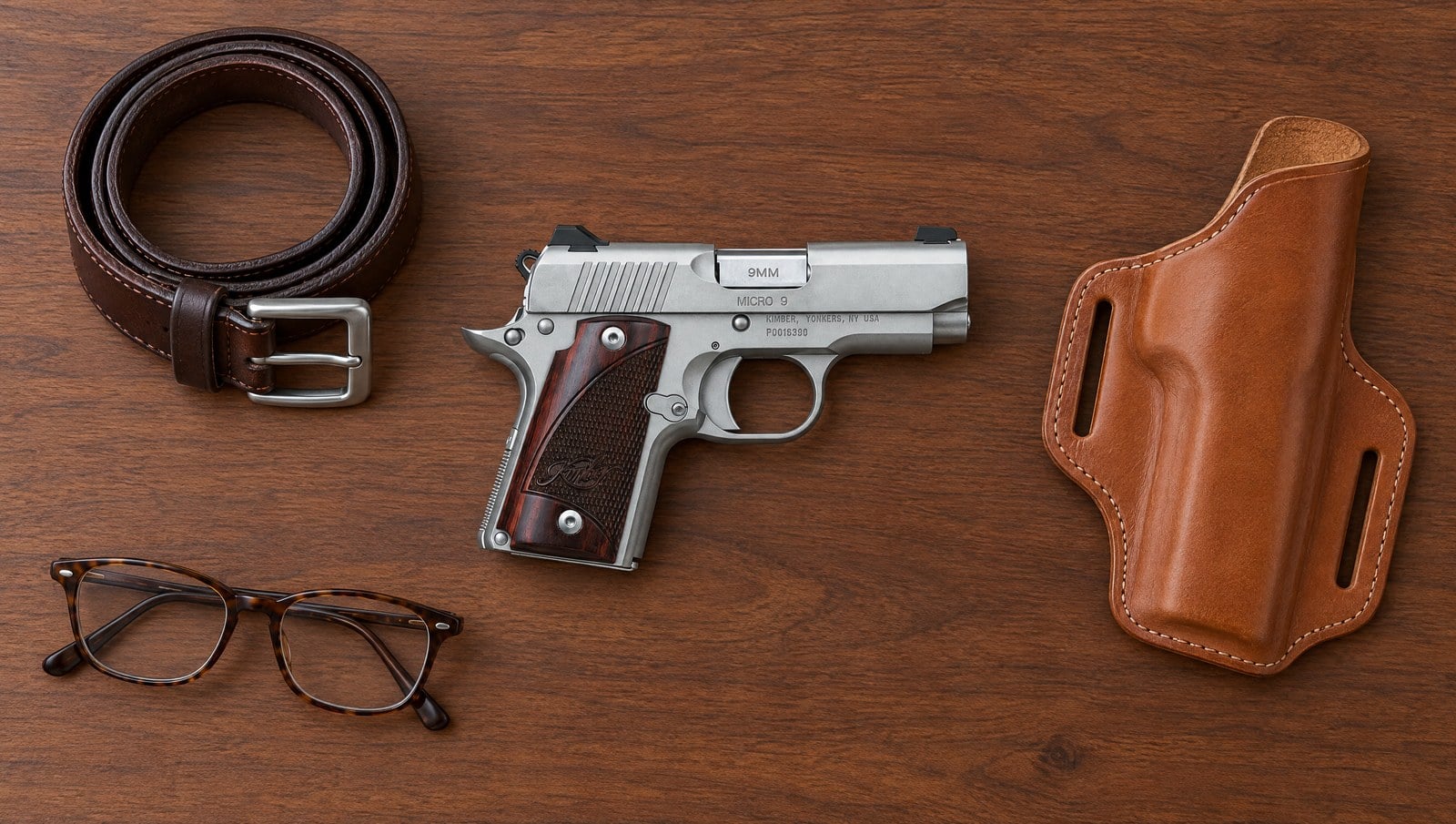 Kimber Micro 9 stainless pistol lying flat on a walnut table beside a tan leather belt holster, a brown belt and reading glasses