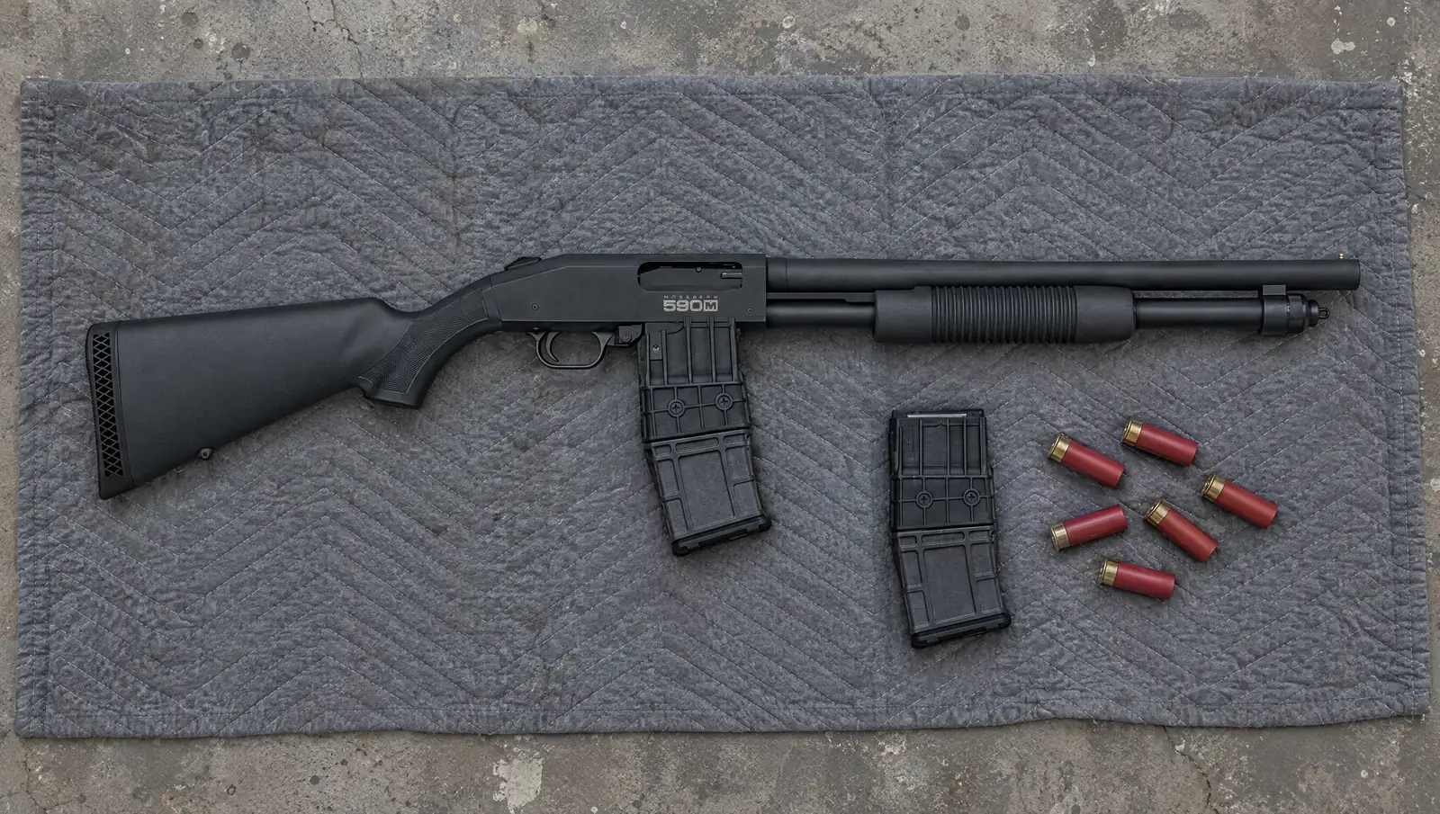 Mossberg 590M Mag-Fed 12 gauge pump shotgun with a 10-round box magazine and bead sight, lying on a moving blanket on a garage floor beside a spare magazine and red shotshells