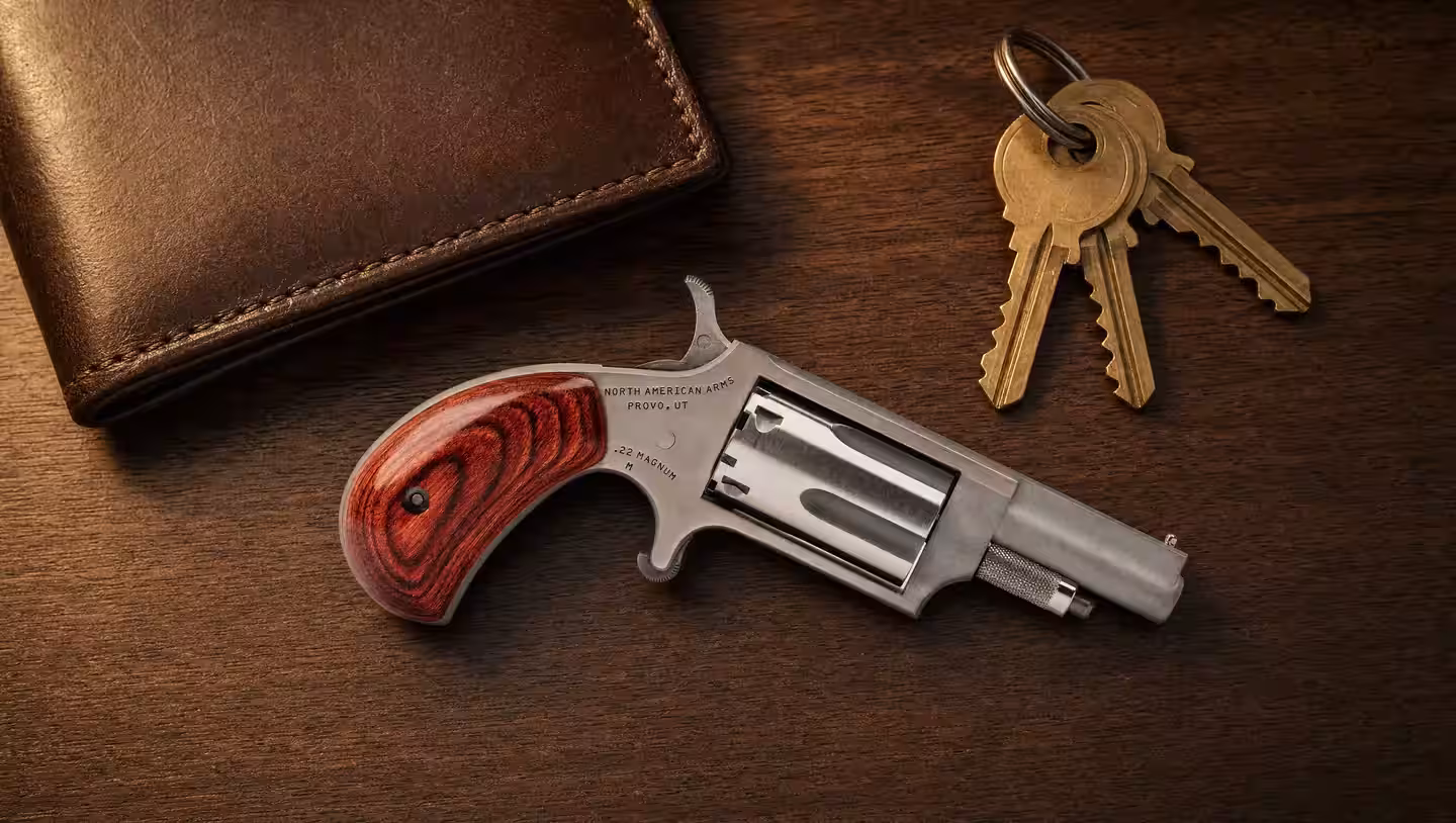 North American Arms Mini Revolver in .22 Magnum with a rosewood bird's head grip lying on a walnut dresser beside a leather wallet and a ring of keys