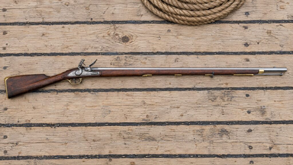Pedersoli Brown Bess flintlock musket lying flat on the pale oak deck planks of a sailing ship beside a coil of rope