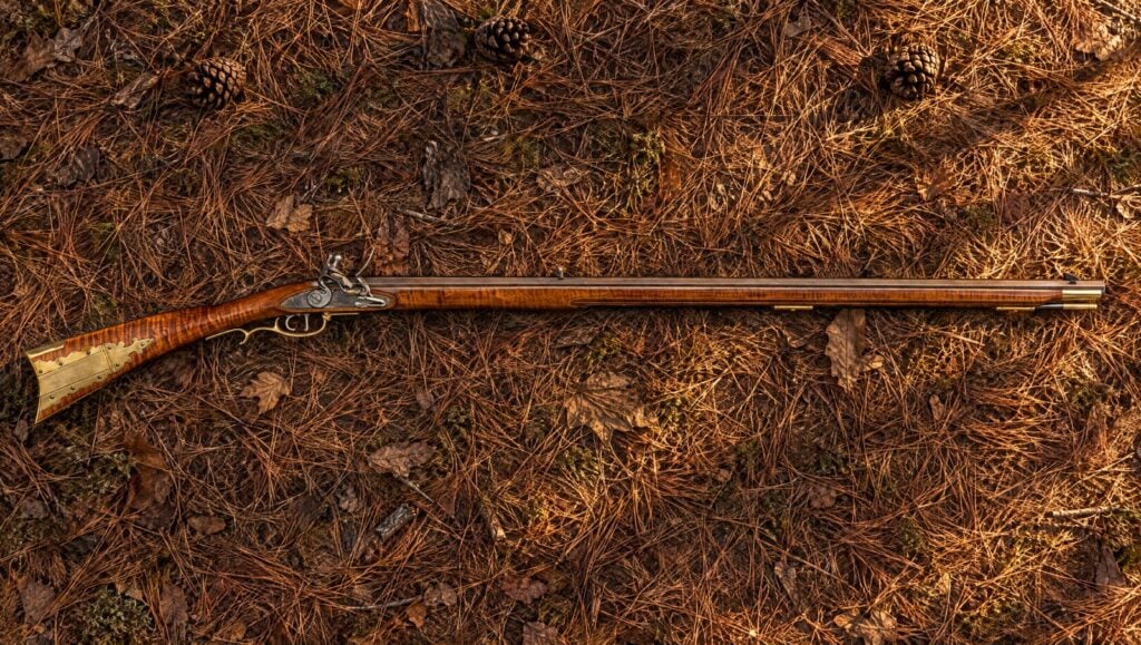 Pedersoli Frontier Maple flintlock long rifle with a striped maple stock lying flat on pine needles in evening sun