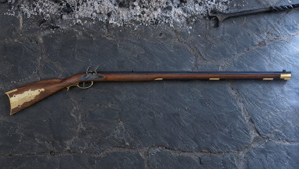 Pedersoli Kentucky flintlock rifle with a large brass patch box lying flat on gray slate hearth stones