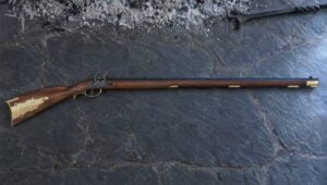Pedersoli Kentucky flintlock rifle with a large brass patch box lying flat on gray slate hearth stones