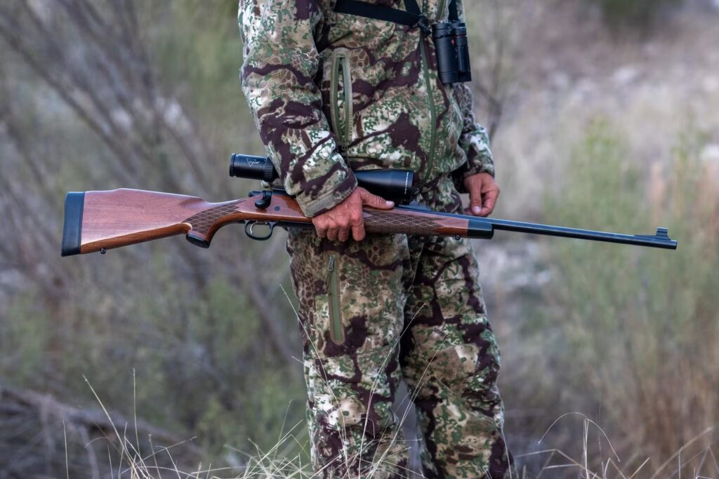 Remington 700 BDL with high-gloss walnut stock, blued barrel, iron sights and a mounted scope held at the hip by a hunter in open brush