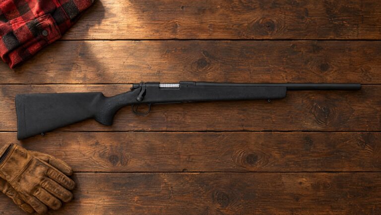 Remington Model 700 bolt-action rifle with a black overmolded stock and heavy barrel lying flat on dark pine floorboards beside a plaid wool shirt