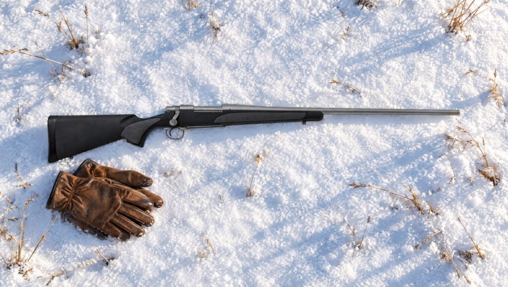 Remington 700 SPS Stainless with matte stainless barreled action and black synthetic stock with gray grip panels lying flat on snow beside leather gloves