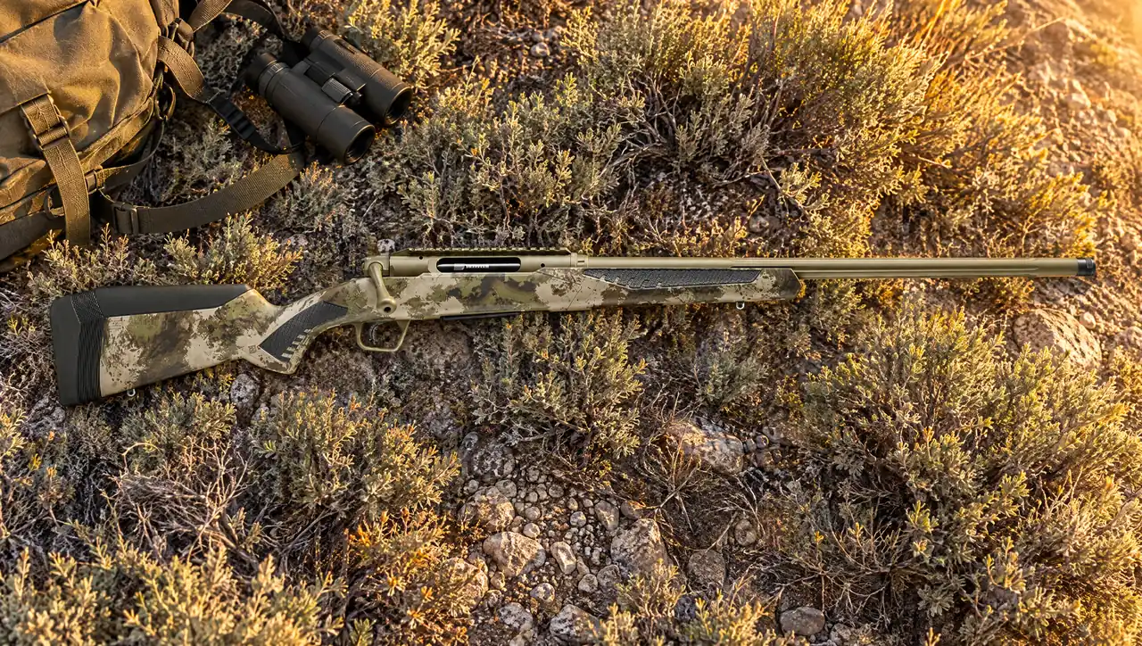 Savage Impulse Big Game straight-pull rifle with Hazel Green Cerakote receiver and fluted barrel and Savage Woodland camo stock lying in sagebrush beside binoculars and a day pack