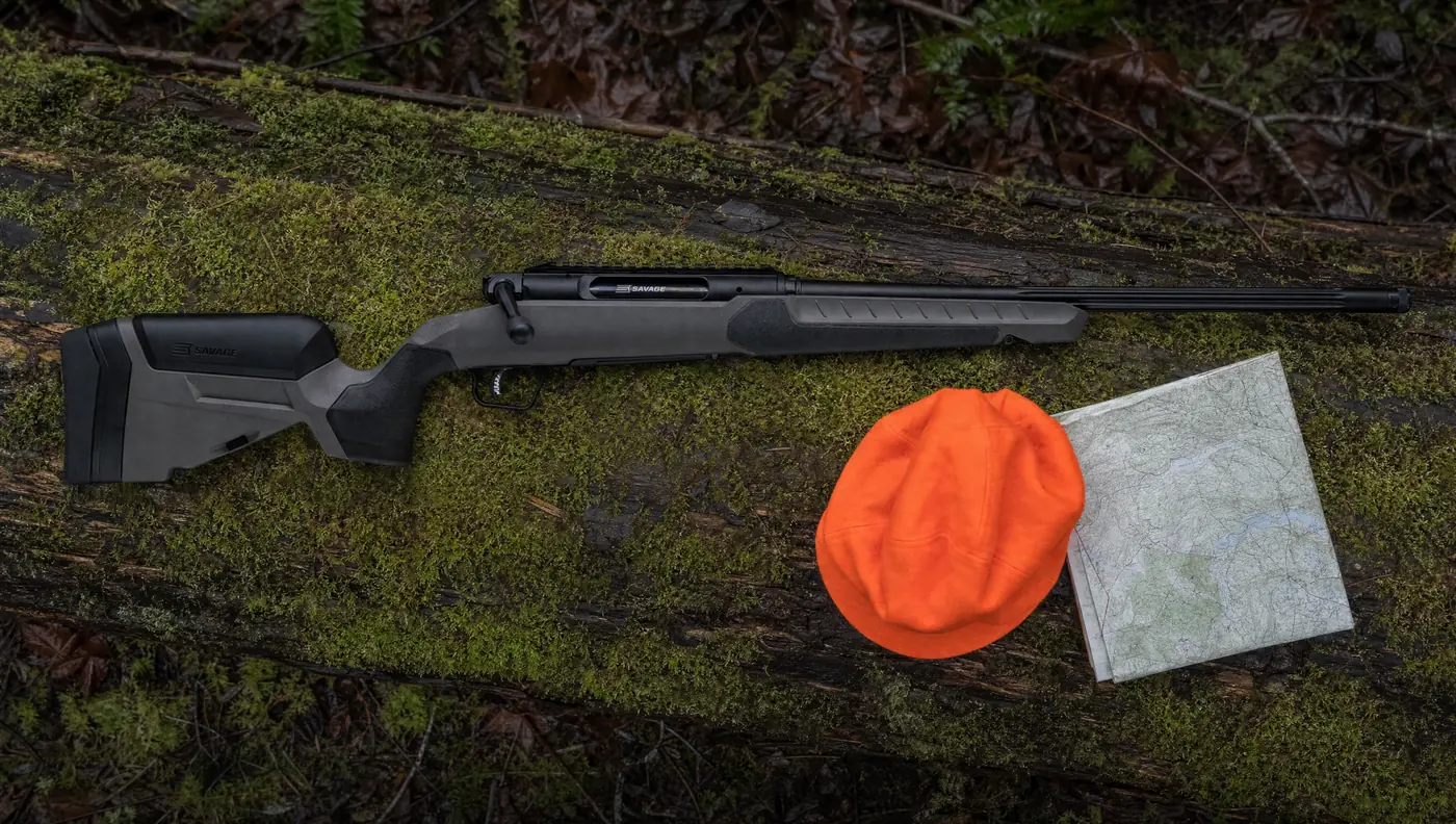Savage Impulse Core Hunter straight-pull rifle with flat dark gray stock and black Cerakote barrel lying on a mossy log beside a blaze orange hat and a map