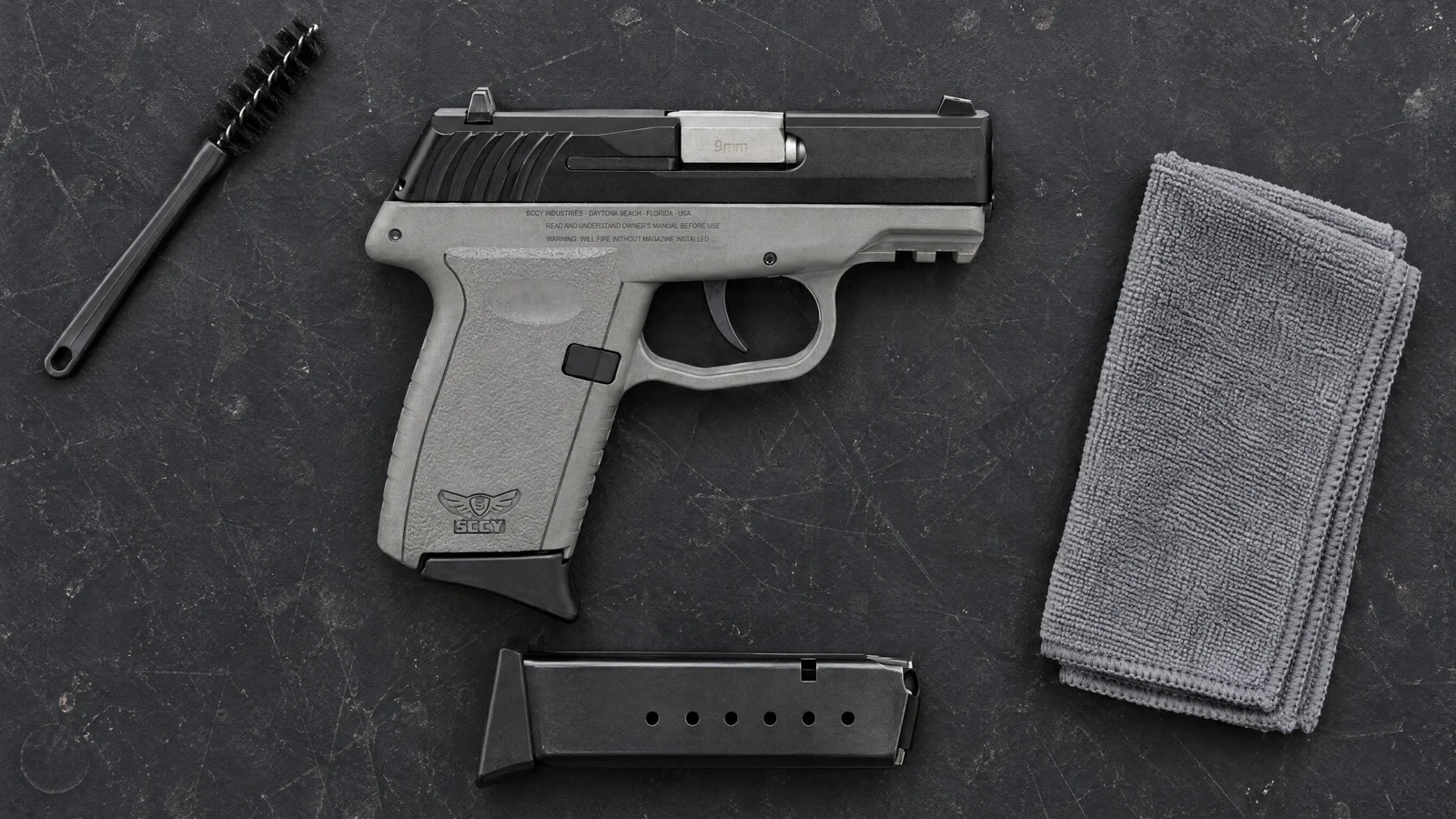 SCCY CPX-2 Gen 3 9mm pistol with a grey frame lying flat on a black workbench mat beside a spare magazine, a cleaning brush and a microfiber cloth