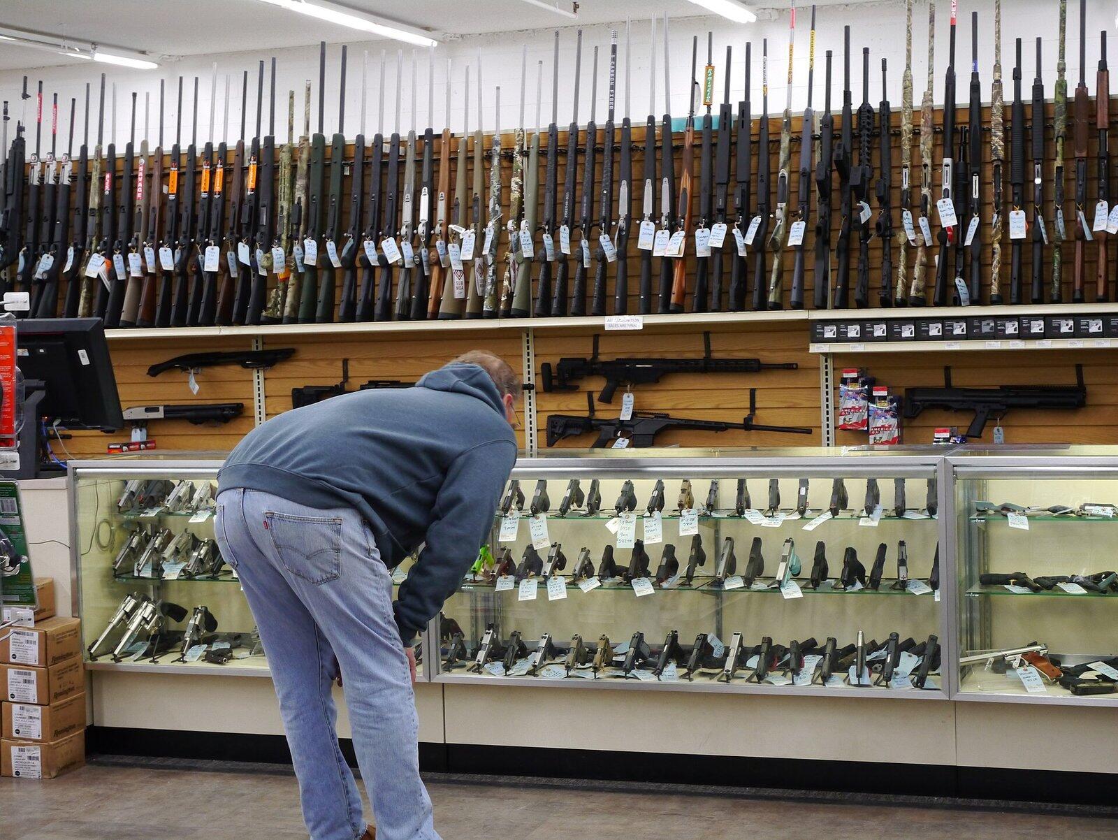 Customer at a glass handgun counter in a gun store, with a wall of tagged rifles behind it
