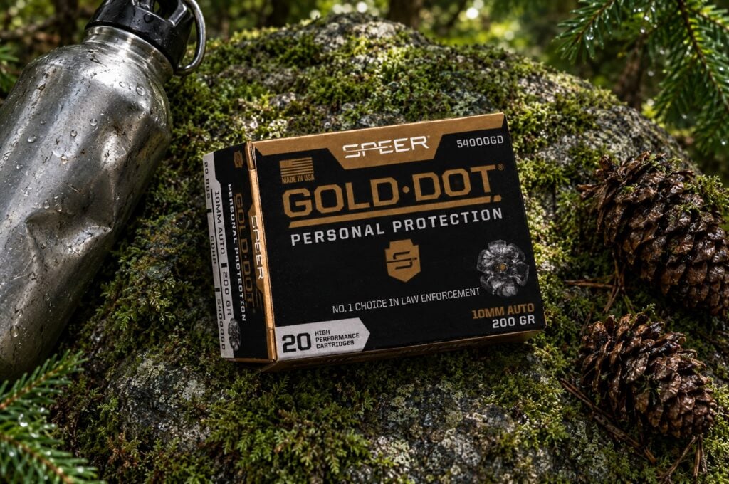 Speer Gold Dot 10mm Auto 200 gr box on a mossy boulder in an evergreen forest beside a steel water bottle