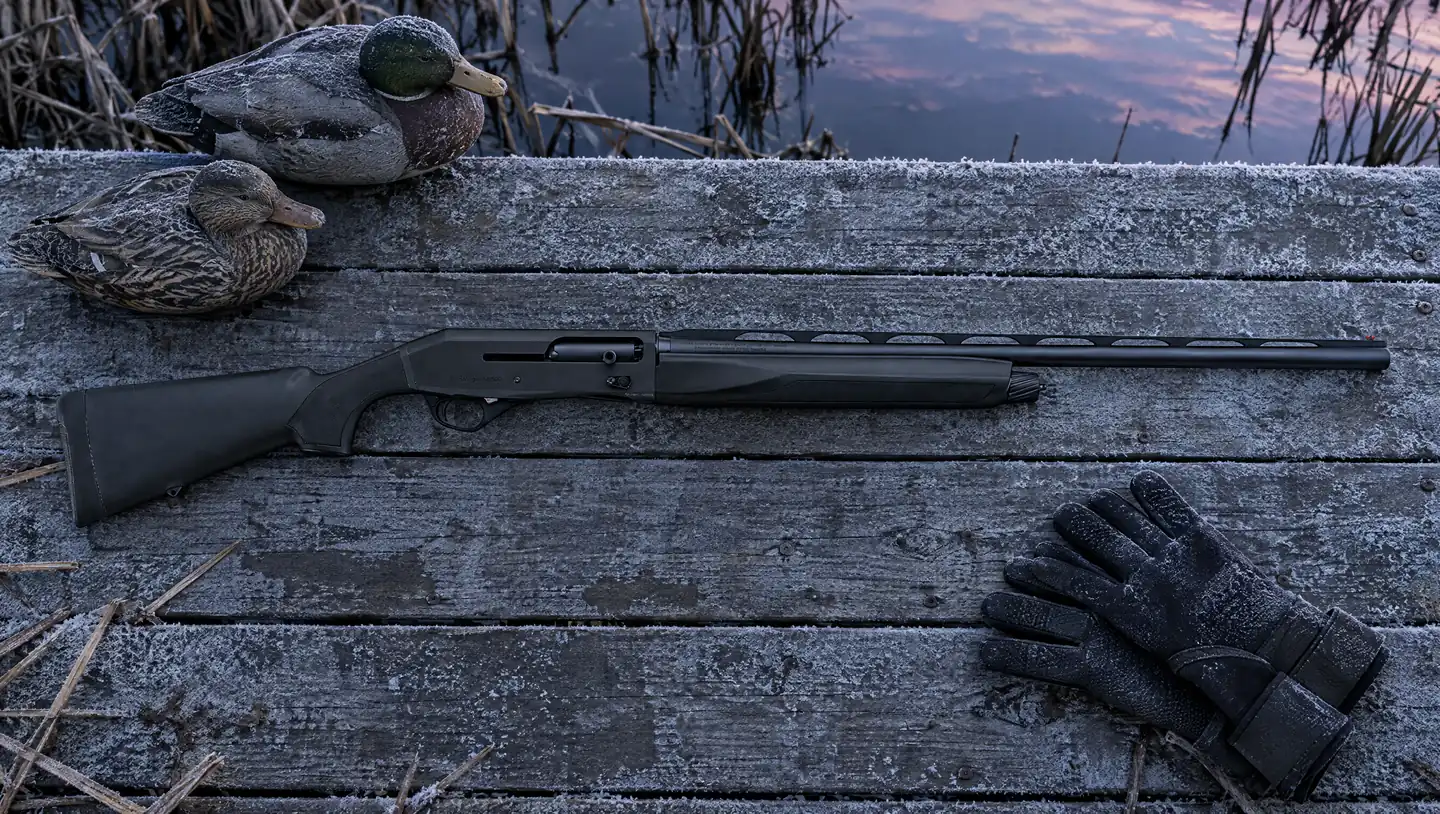 Black synthetic Stoeger M3500 12 gauge semi-auto shotgun lying on a frosty duck boat dock at dawn beside mallard decoys and neoprene gloves