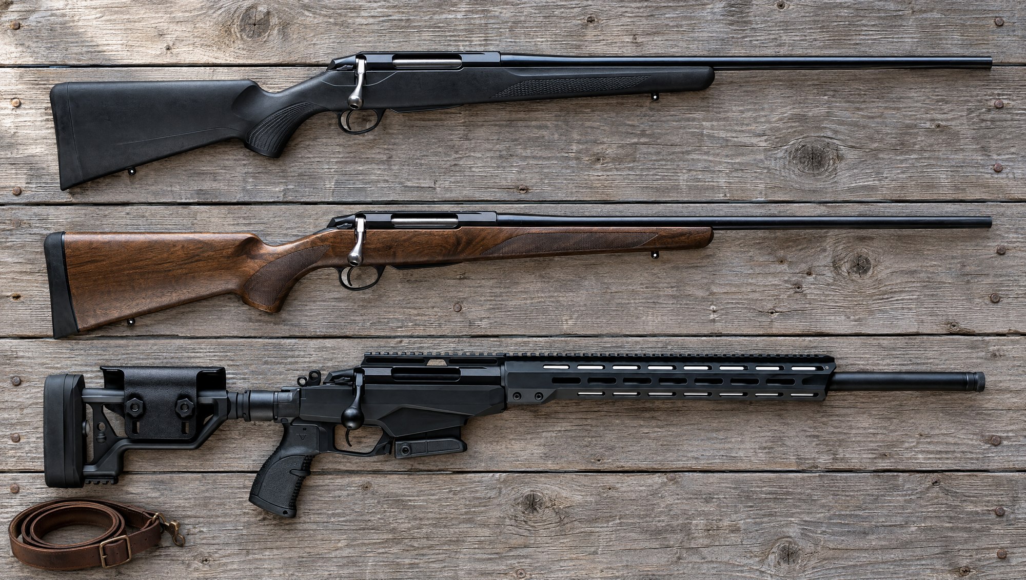 Tikka T3x models compared: a black synthetic T3x Lite, a walnut T3x Hunter and a T3x TAC A1 chassis rifle lying flat on barn wood