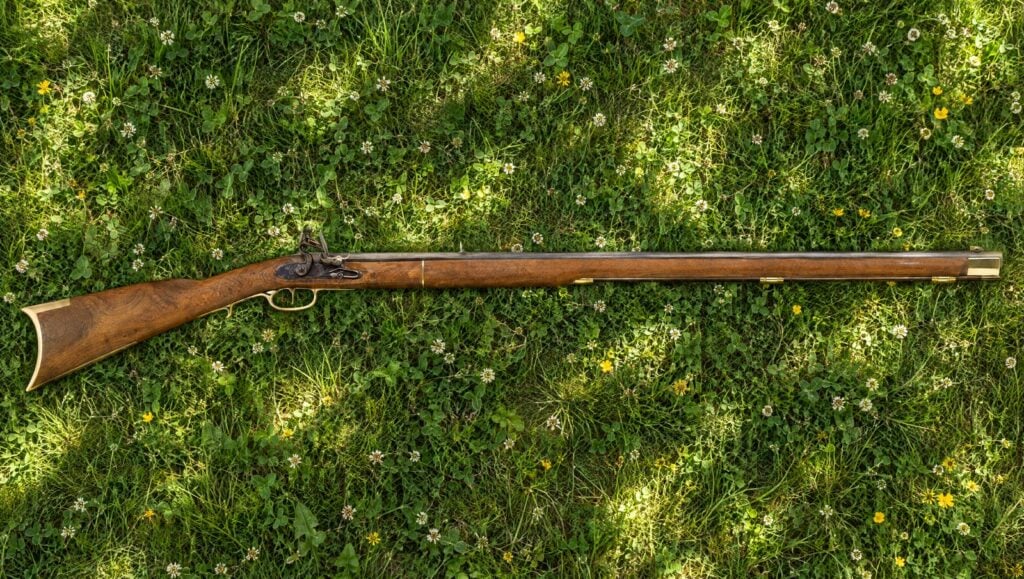 Traditions Kentucky flintlock rifle lying flat on meadow grass with clover in dappled shade