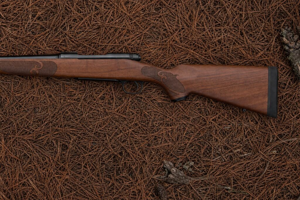 Winchester Model 70 Featherweight left side close-up on pine needles, showing the walnut buttstock, grip checkering, trigger guard and recoil pad